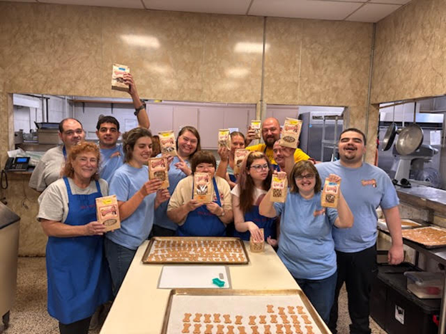 Group of people in a kitchen holding up packages, likely in a celebratory or team-building moment.