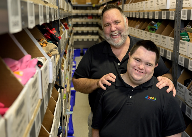 Two men standing in a storage area with shelves filled with boxes and items.