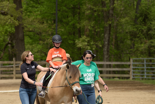 Two women guiding a child on a horse in an outdoor setting with trees in the background